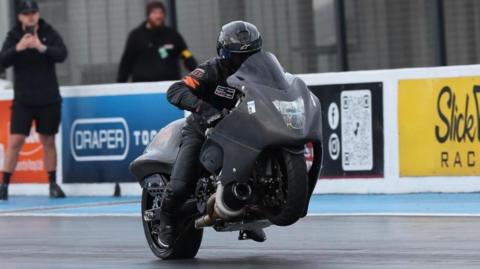 A Suzuki Hayabusa Turbo drag bike speeding along the track at Santa Pod. It is black with a large protective shield at the front, and Dan is clad head to toe in black protective clothing, and a black helmet. Two men stand at the side, one possibly taking a photo with his phone. There are adverts on the standings to one side. 