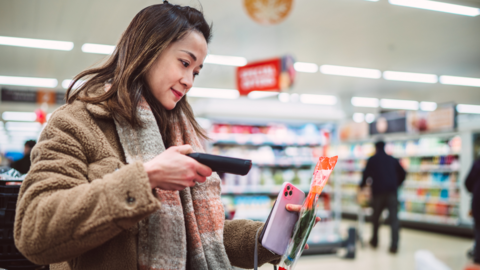 Young woman with dark hair, wearing brown fake fur jacket and grey and pink scarf scans packet of herbs at supermarket  