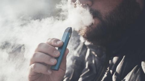 A bearded man surrounded by smoke holds a blue vape to his mouth