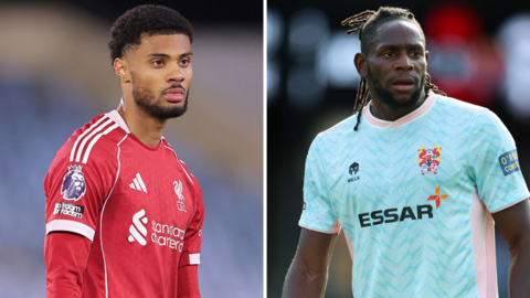 Kyle Kelly standing on a pitch wearing red Liverpool kit and Omari Patrick (right) standing looking to his left wearing light blue Tranmere Rovers kit during a match