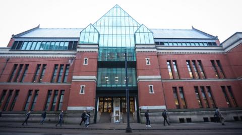 A general view of Minshull Street Crown Court in Manchester. The court is a two-storey red brick building with a glass-panel frontage. People can be seen walking past the entrance on a grey day. 