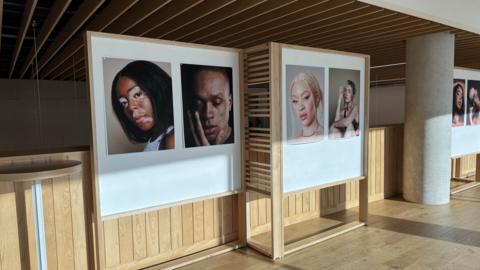 The exhibition of 12 portraits of models' faces has been set up in a university gallery. There are pairs of images on white boards around a wooden panelled room.