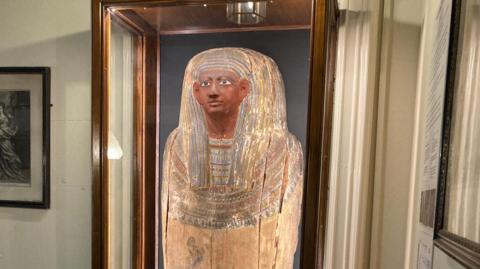 An ancient Egyptian coffin lid which depicts the effigy of a person. It is yellow, brown and blue in colour.