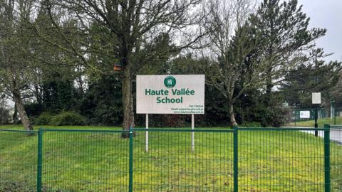 A sign for Haute Vallée School. It is green and white. The sign is behind a large green metal mesh fence and is on a grass bank. Several trees and bushes are behind it.