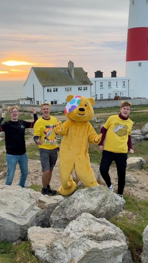 Pudsey is a yellow fluffy human sized bear with a white eye patch covered in green, purple and red polka dots. He stands with Steve Harris on his right and two other BBC producers, who are all wearing yellow Children in Need t shirts in front of a red and white striped lighthouse as the sun rises. They are raising their arms as if they are cheering.