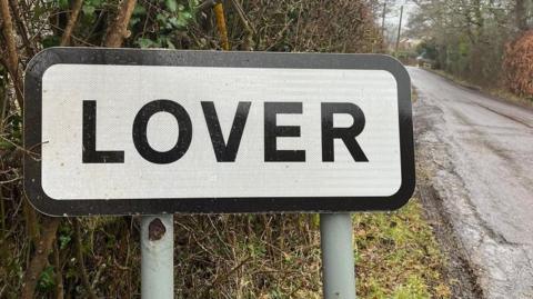 The Lover road sign on a wet day next to a hedge and road.