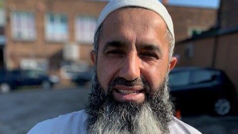 A bearded man wearing a cream taqiyah. He is slightly smiling and the picture was taken in a car park