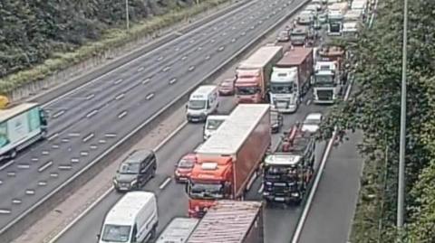 Queuing traffic on the M25 . There are a mix of lorries, vans and cars tightly packed across three lanes of traffic on the road.