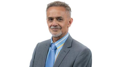 Ranjit Gill, a man with short grey hair and a short grey goatee beard. He has a straight facial expression. He is wearing a grey suit, a light blue shirt with white and yellow stripes, and a slightly darker blue tie.