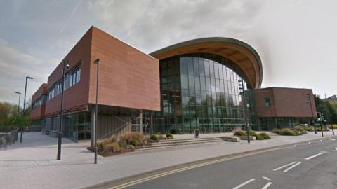 A large, glass-front building at the University of Warwick. The pavement and road in front of the building are also visible, and bushes line the steps and entrances to the building.