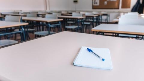 A stock picture of an empty classroom, with a notepad and blue biro on top of it.
