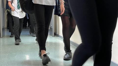 Unidentified high school children walking in a school corridor. The floor is a grey colour. The pupils are wearing black shoes and one of them has a green and white stripey tie, with one in a green tartan skirt. 