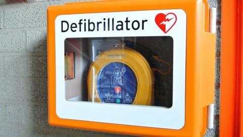 An orange defibrillator box with a glass front and orange circular bag inside. There is a diagram on the front of the bag showing how to use the device.