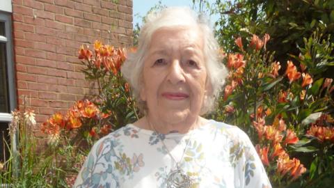 Head and shoulders image of Phyll Babb, wearing a white top and standing in front of some orange flowers