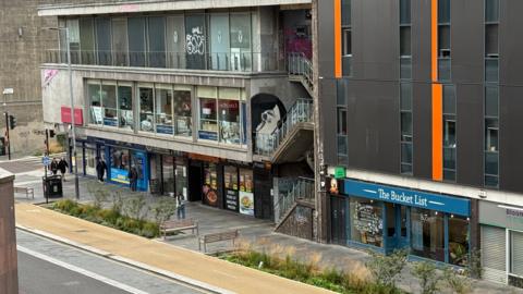 Retail and hospitality venues in buildings of mixed architectural styles line Cambridge Street, which has roadwork bollards along the middle of the road. 