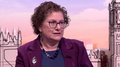 Hilary Cass on the Sunday With Laura Kuenssberg set. She has medium-length curly brown hair and is wearing a purple jacket, dark blue top and has a large purple bead necklace and purple bead earrings.