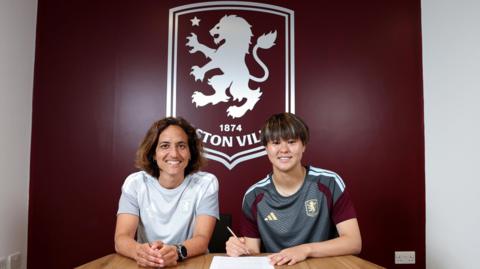Akane Okuma signs contract in front of large Aston Villa emblem