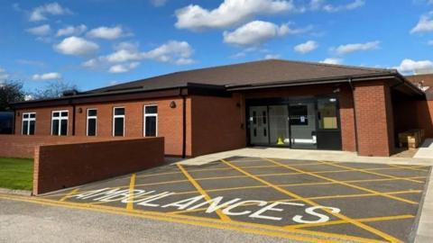 The outside of a single-storey, red brick building with a bay at the front. It has yellow cross-hatched lines and "Ambulance" spelled out in white capital letters, indicating a drop-off zone.