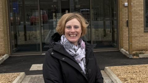 A woman with light brown hair wearing a dark coat and scarf standing outside a building with large glass doors.