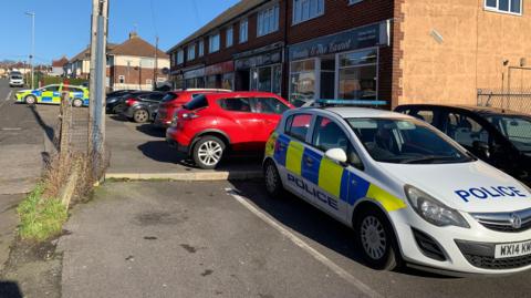 Two police cars are parked up outside a parade of shops with flats above them. There are other cars also parked up along the street, with houses visible in the background.