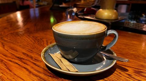 An image of a grey coffee cup with frothy oat milk. Two packets of brown sugar and a silver coloured spoon are on the saucer 