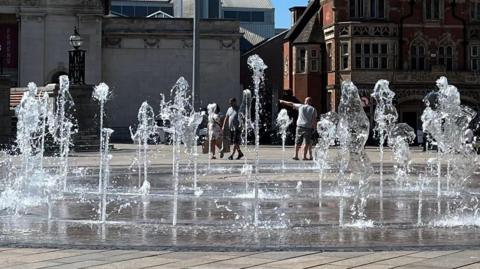 Columns of water bubble up from beneath a pavement in a city square, with early 20th century buildings in the background and several people walking past.