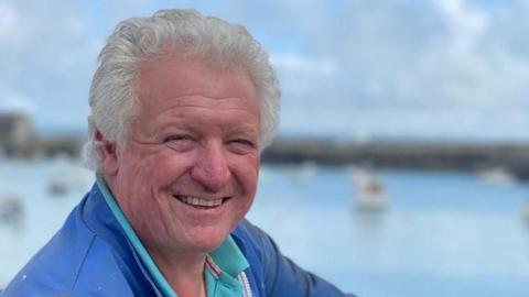 A man smiling at the camera wearing a blue shirt and blue jacket with the background of the ocean blurred.