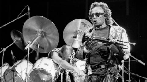 An African American man wearing a jacket, t shirt and sunglassesholding a trumpet. He is standing in front of a set of drums. There is a man behind the drums leaning forward over them. It could be a shot taked from a performance.