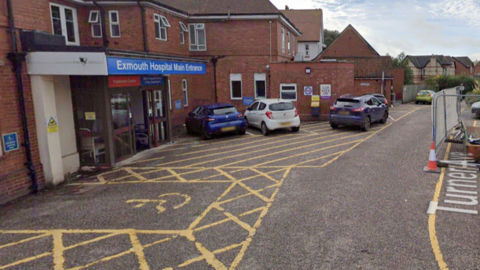 A street-view of Exmouth Hospital main entrance. There are yellow disabled parking bays outside of the entrance with cars parked on the other side. The sign at the top of the entrance is blue.  