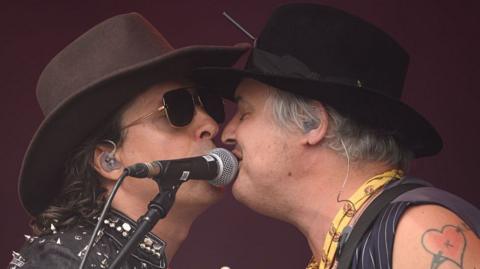 Live performance energy, captured mid-connection. This image shows Carl Barat and Pete Doherty of the Libertines locked in a moment of animated exchange on stage. Peter (right)—wearing a black hat, sleeveless top, and a yellow patterned scarf—has a tattoo of a heart with an arrow on their upper arm. Carl (left) is dressed in a brown hat and a studded leather jacket, exuding classic rock ’n’ roll vibes. Both are equipped with in-ear monitors