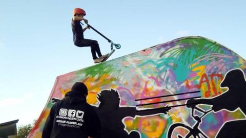 The image shows a young person on a scooter on the top of a ramp.  The ramp has been decorated in bright stree art, with images of black cutouts of a skater and BMX rider.
