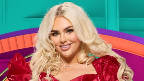 Jenny Baird has long blonde hair and is standing in a red dress. Her hands are on her hips as she stands in front of a large colourful eye (the Big Brother logo).