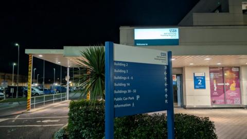 A blue and white sign with labels and arrows. A hospital building is in the background and a car park is to the left. It is night time.