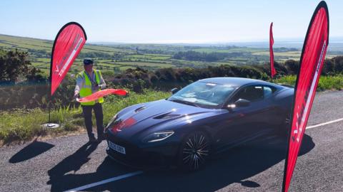 A sports car drives along a scenic country road. There are flags on either side of the road and a man in a yellow high-vis vest waving a red flag.