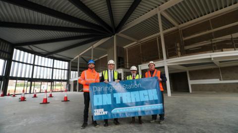 Luke Durston (Network Rail), Tees Valley Mayor Ben Houchen, councillor Steve Harker and Steve Wilson of LNER hold a blue banner reading Darlington Station On Track, with a train on it, inside the new concourse. It is a large circle building with panels on the ceiling and tall windows.