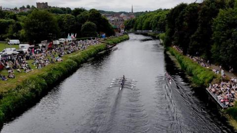 An aerial view of the Durham Regatta. Two rowing boats are on the River with corwds of people watching from the banks. The skyline of Durham City is in the background.