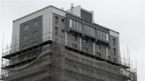 The top of St Francis Tower. Gray and white cladding is visible at the top few floors with scaffolding and netting on lower floors. 