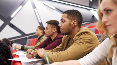 A stock photo shows four students sat in a row of a lecture hall, as taken from the side. Three are men and there is a young woman in the foreground. They have papers in front of them and pens in their hands, and are looking intently to the left, towards the lecturer at the front.