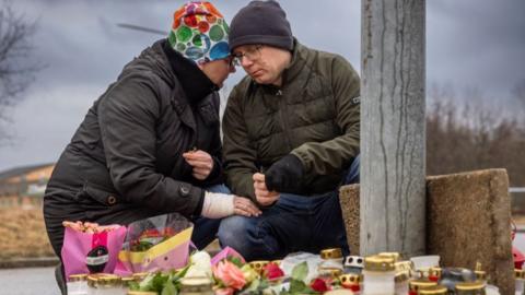 Two people touch their heads and lean over candles and flowers at a memorial