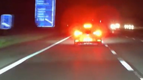 A car's rear brake lights are illuminating in red on lane one of a motorway. To the left are blue motorway signs. 