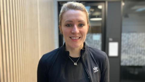 A woman with blonde hair, a black Adidas jacket, black top and silver necklace, stands and smiles. 