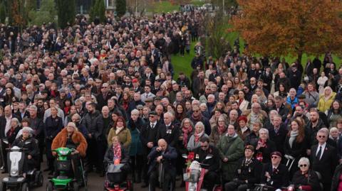 Military veterans and guests during the Remembrance Sunday service at the National Memorial Arboretum. There are hundreds of people in a line. Some sit on mobility scooters at the front. Others are standing. Many are wearing suits and poppies. One man can be seen wiping his eyes. 