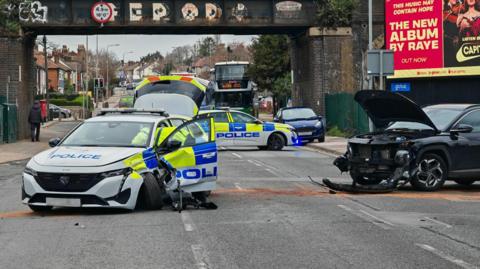 A police car is on the left of the image, its front left wheel is damaged and there is damage to the door and front of the vehicle. On the right of the picture is a black car, its bonnet is lifted with damage to the front bumper. A police car in the background is blocking off the road.
