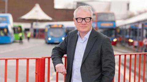 Richard Parker wearing black glasses, a grey suit leaning on a red fence in front of a bus stop 