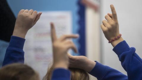 A classroom. Children in blue jumpers put up their hands to answer a question.