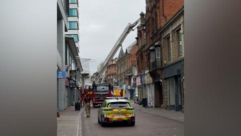 A police car, fire engine and ambulance are parked on a pedestrian street. A firefighter in blue t-shirt with yellow firefighting trousers is walking on the left hand side of the road. There is a crane appliance extended up to a roof.