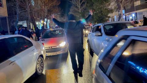 A person walks between moving cars on a road, while making the victory sign with his hands, at night-time in Hamedan, Iran (01/01/26)