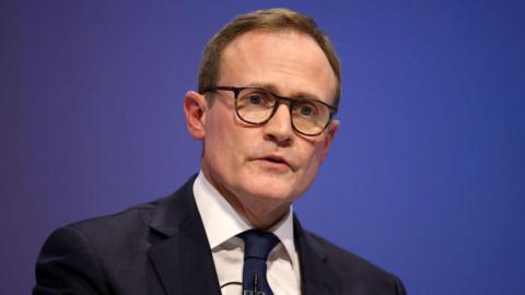 A man wearing glasses and a dark suit, white shirt and dark blue tie. 