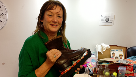 Sandra Leckey, in green sweater, holding up brown boots