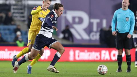 Alfie Massey runs with the ball in the dark blue and white shirt of Millwall, pursued by a defender in a yellow shirt for Dagenham & Redbridge as a referee in blue looks on in the background.